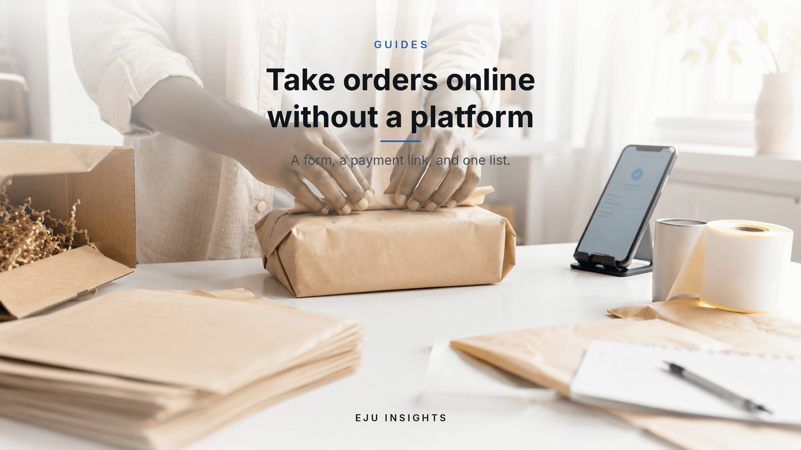 Someone wrapping a parcel in brown paper at a bright white table, with a phone showing an order confirmation and packing envelopes beside it.